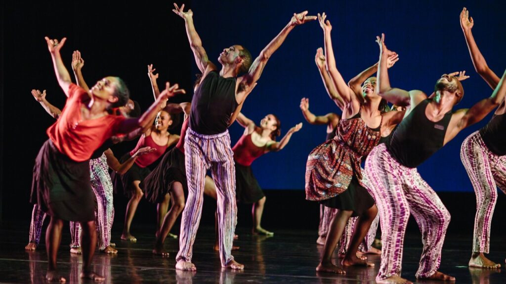 Several dancers in patterned clothing and bare feet hold their arms high and smile on a black stage.