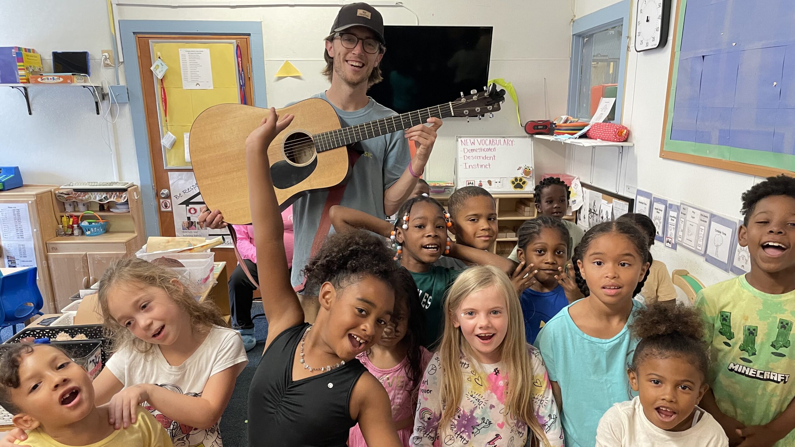 Thirteen young kids smile and pose in front of an adult holding a guitar in a classroom.