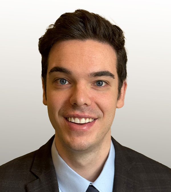 Cameron is smiling, wearing a charcoal gray suit and a light blue shirt. He has dark brown hair and blue eyes.
