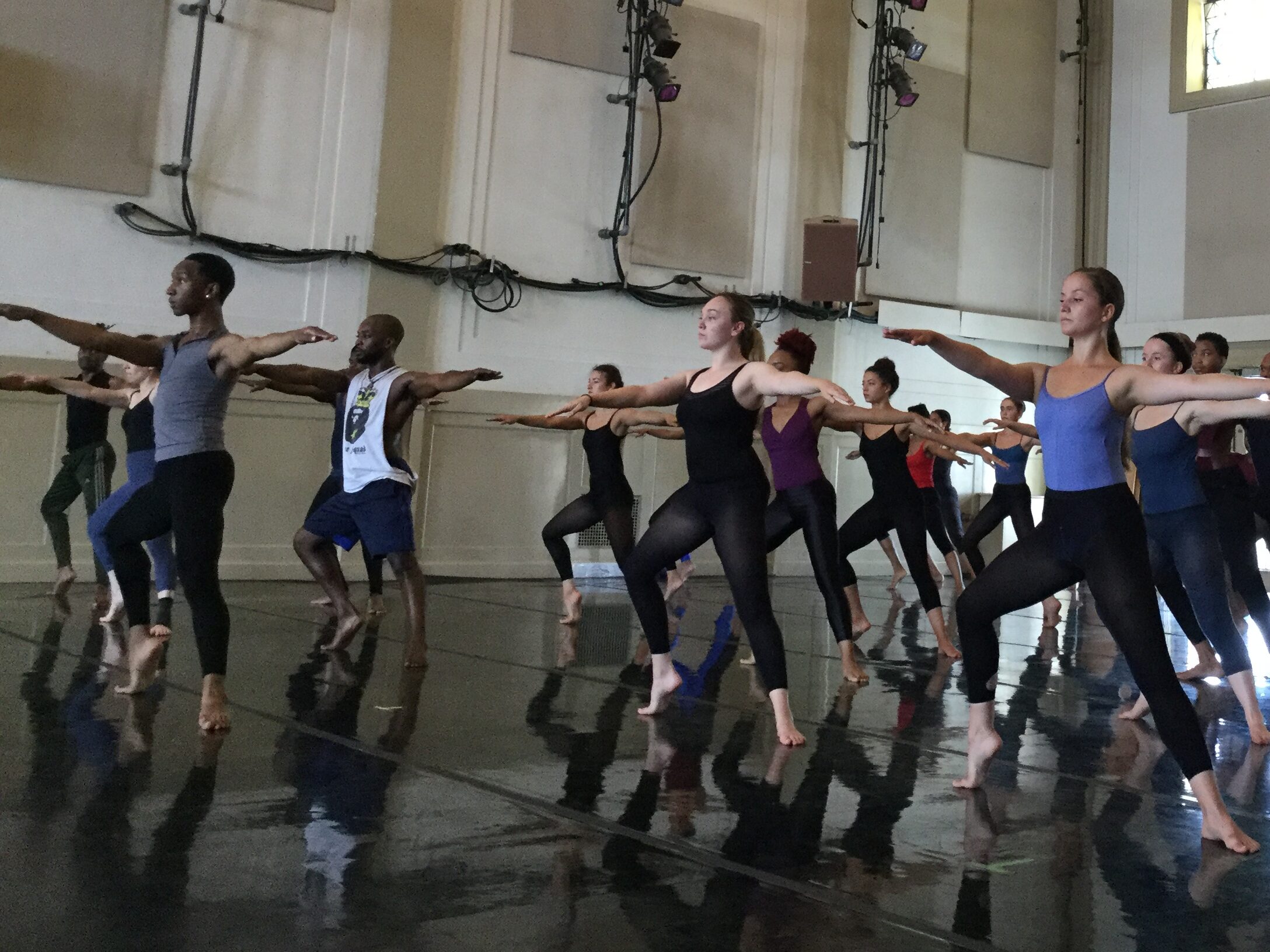 Dozens of dancers in bare feet practice on pointed toes in a ballroom.