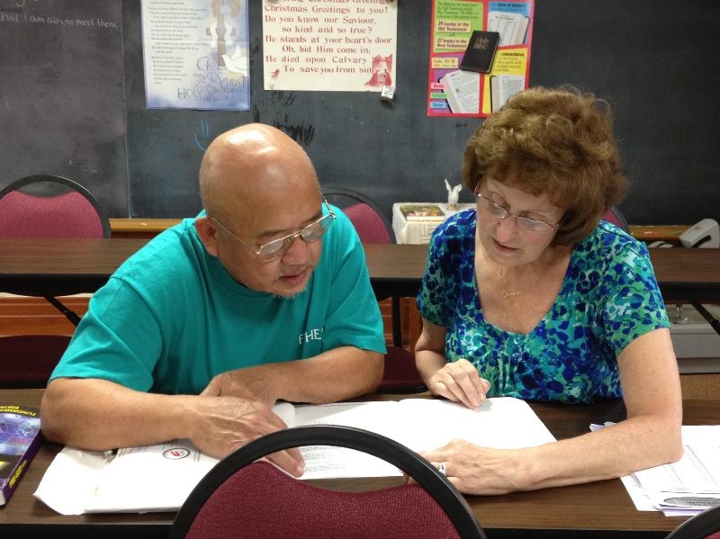 A man in a teal shirt and glasses and a woman read papers together at a desk.