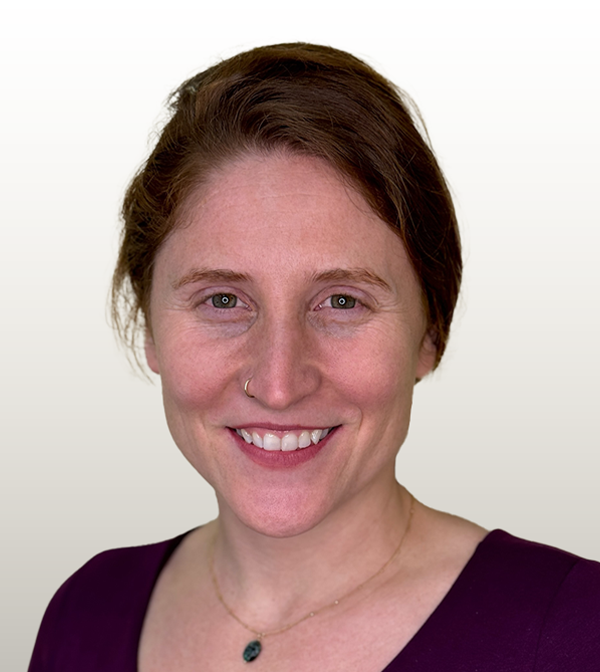 Sarah has red hair in a ponytail and is smiling while wearing a purple shirt.
