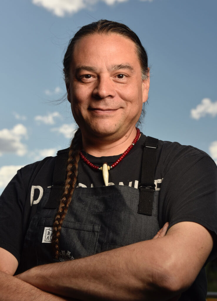 Sean Sherman stands with his arms crossed wearing a black chef's apron and a red beaded necklace. His long braid rests over his right shoulder.