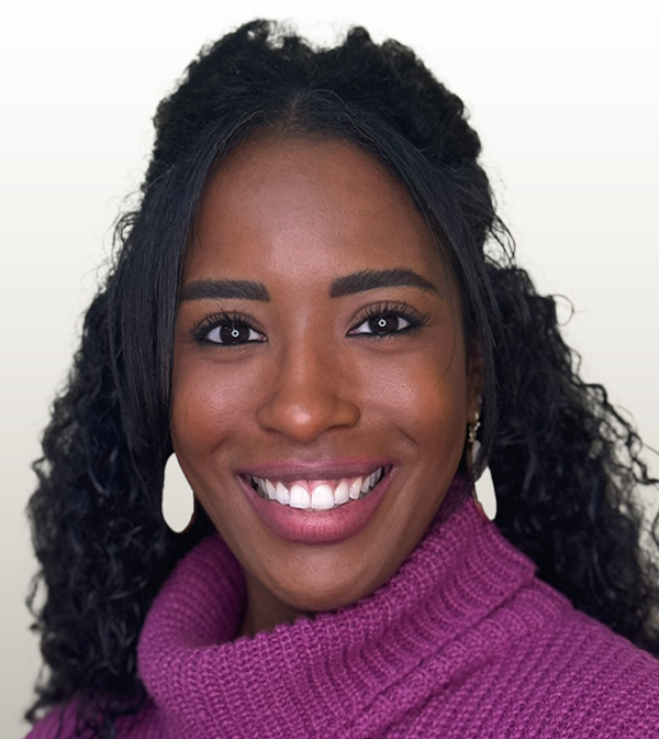 Tyana is smiling in a purple turtle-neck. Her dark hair is long and she is wearing white earrings.