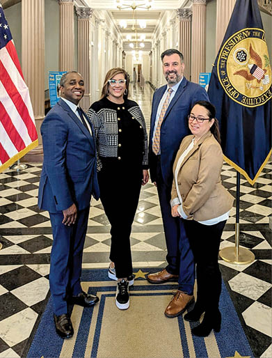 Four people in suits pose in an elegant hallway next to the US flag.
