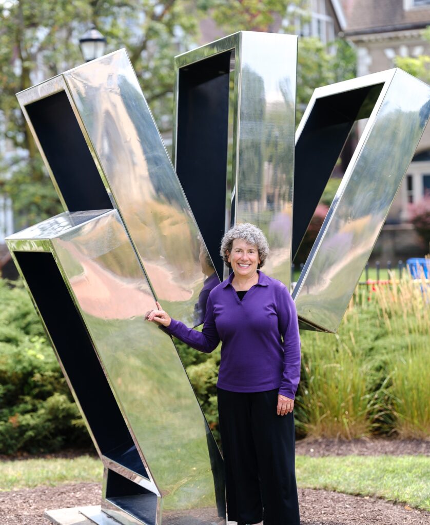 A woman in a purple shirt stands in front of an outdoor sculpture.