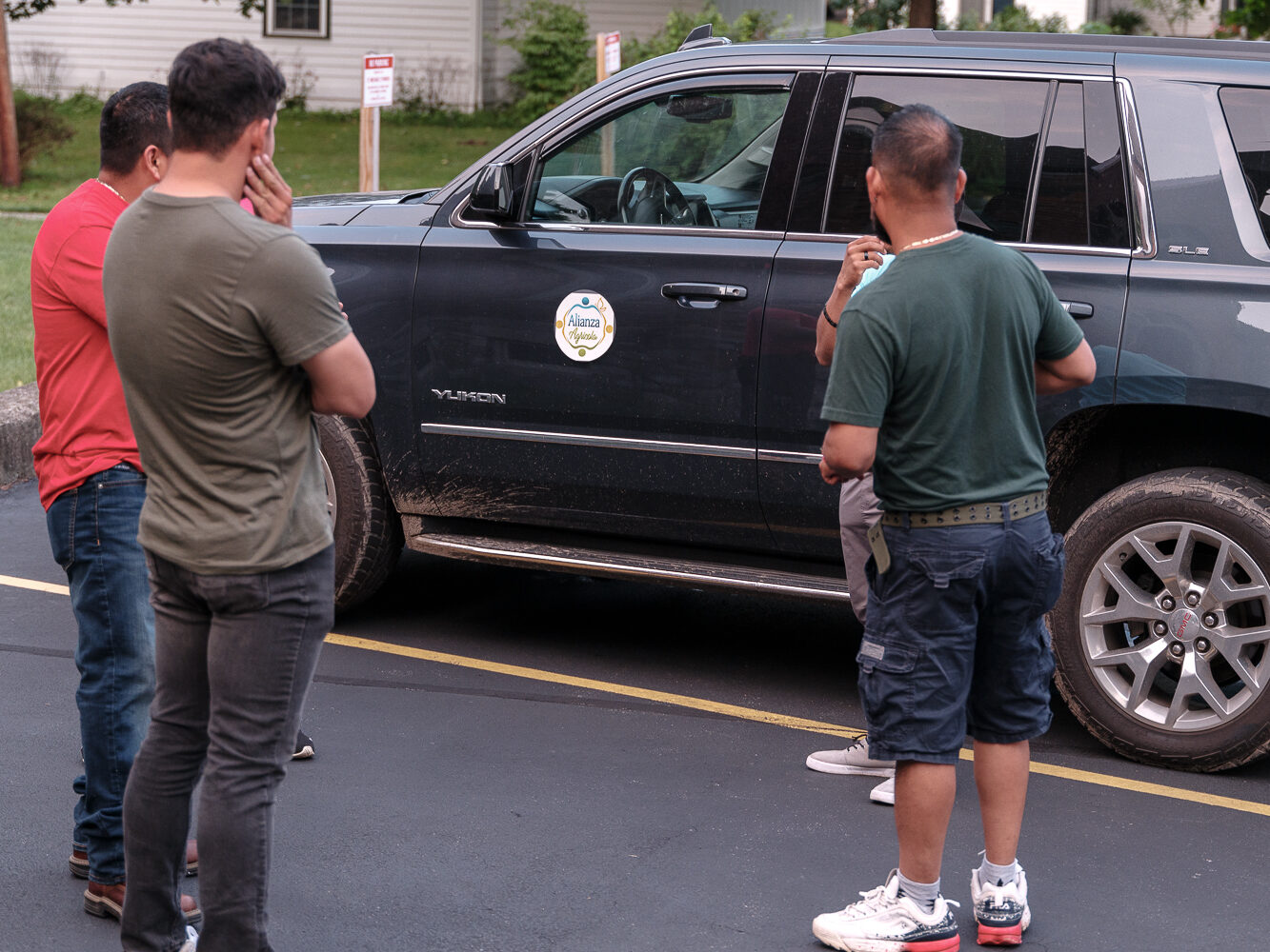 People in sneakers stand casually in front of a black car with mud on its frame and an Alianza Agricola emblem on the door.