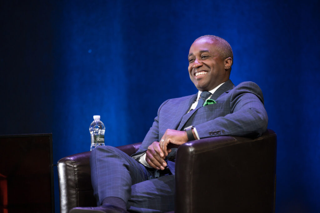 A man in a blue suit sits in an armchair in front of a royal blue background