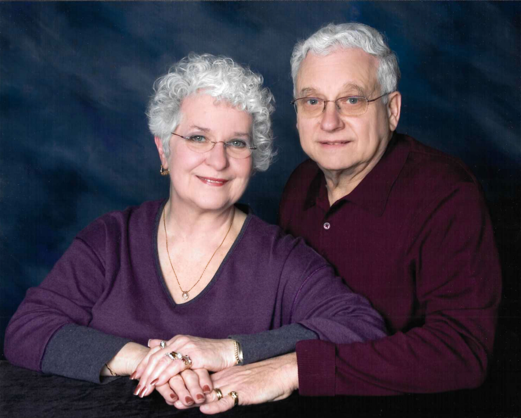 A portrait of a women in a purple sweater and gold jewelry smiling next to a man in a maroon button-down.