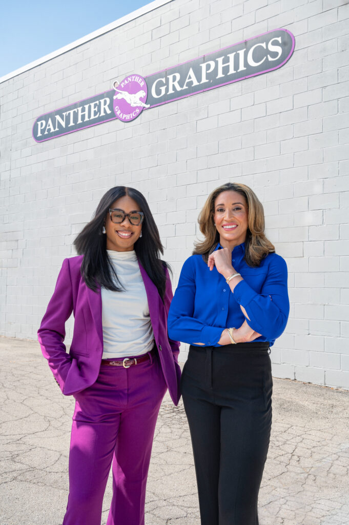 A woman in a purple suit and glasses stands with hands in her pockets next to a woman in a blue shirt and black pants in front of a Panther Graphics sign.