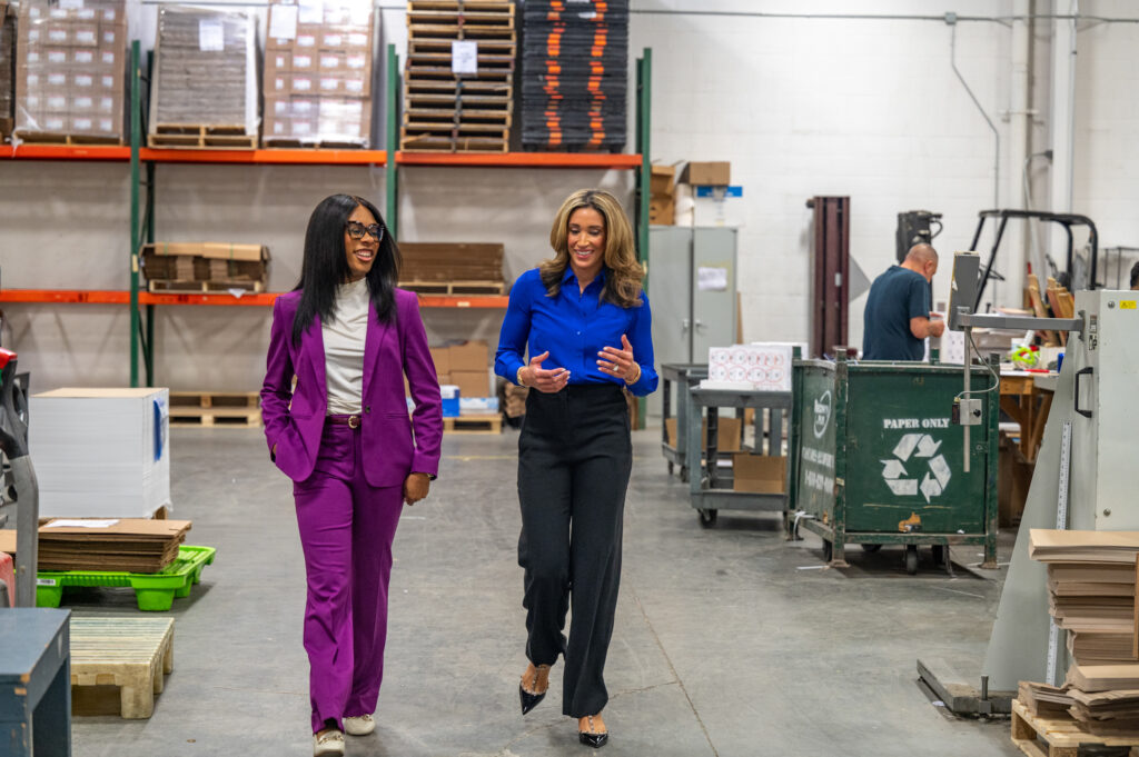 A woman in a purple suit and glasses walks through a warehouse with one hand in her pockets next to a woman in a blue shirt and black pants.