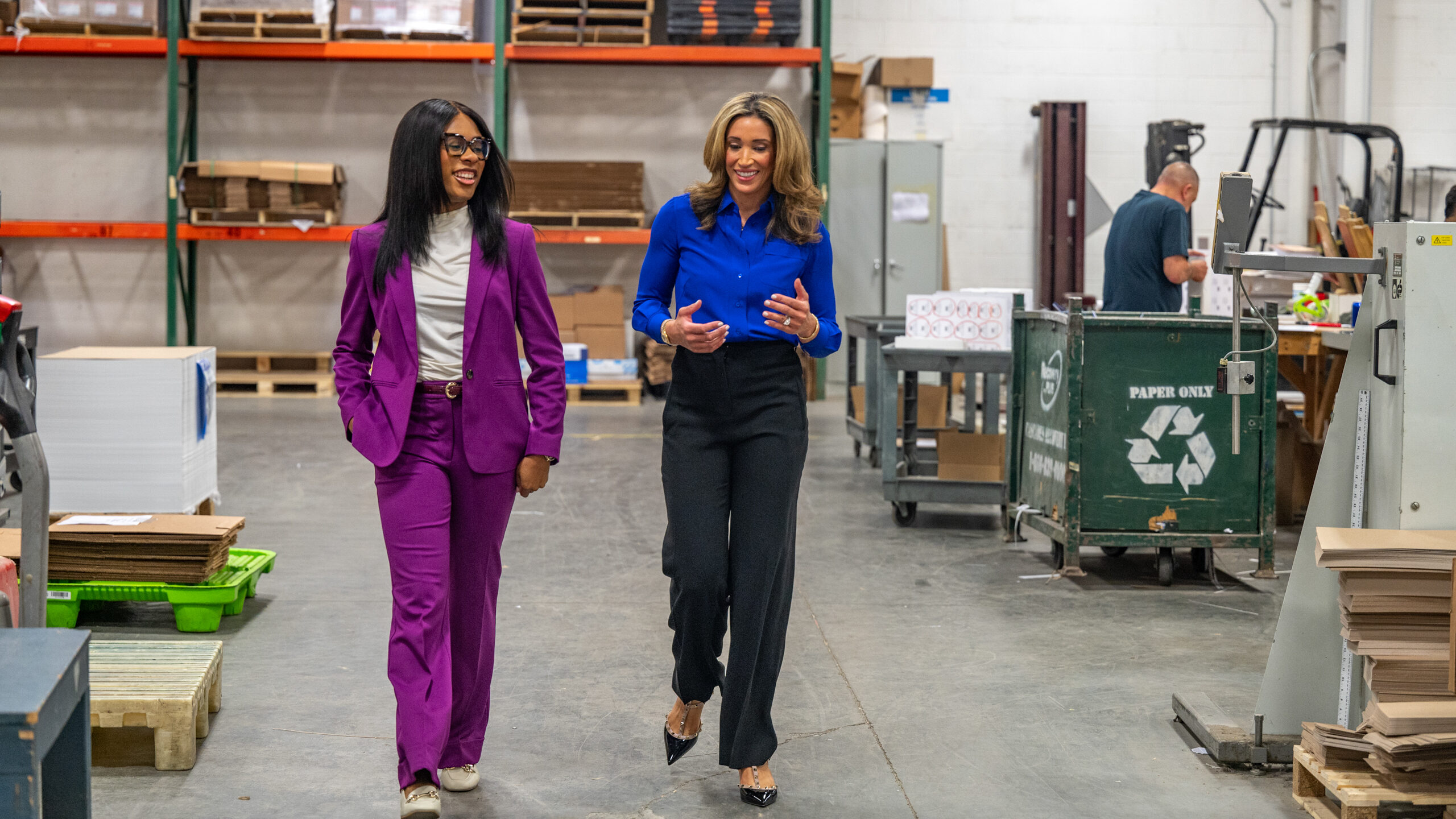 A woman in a purple suit and glasses walks through a warehouse with one hand in her pockets next to a woman in a blue shirt and black pants.