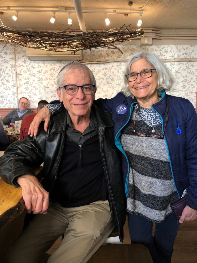A woman in glasses and a striped shirt has her arm around a man in glasses and a leather jacket in a restaurant