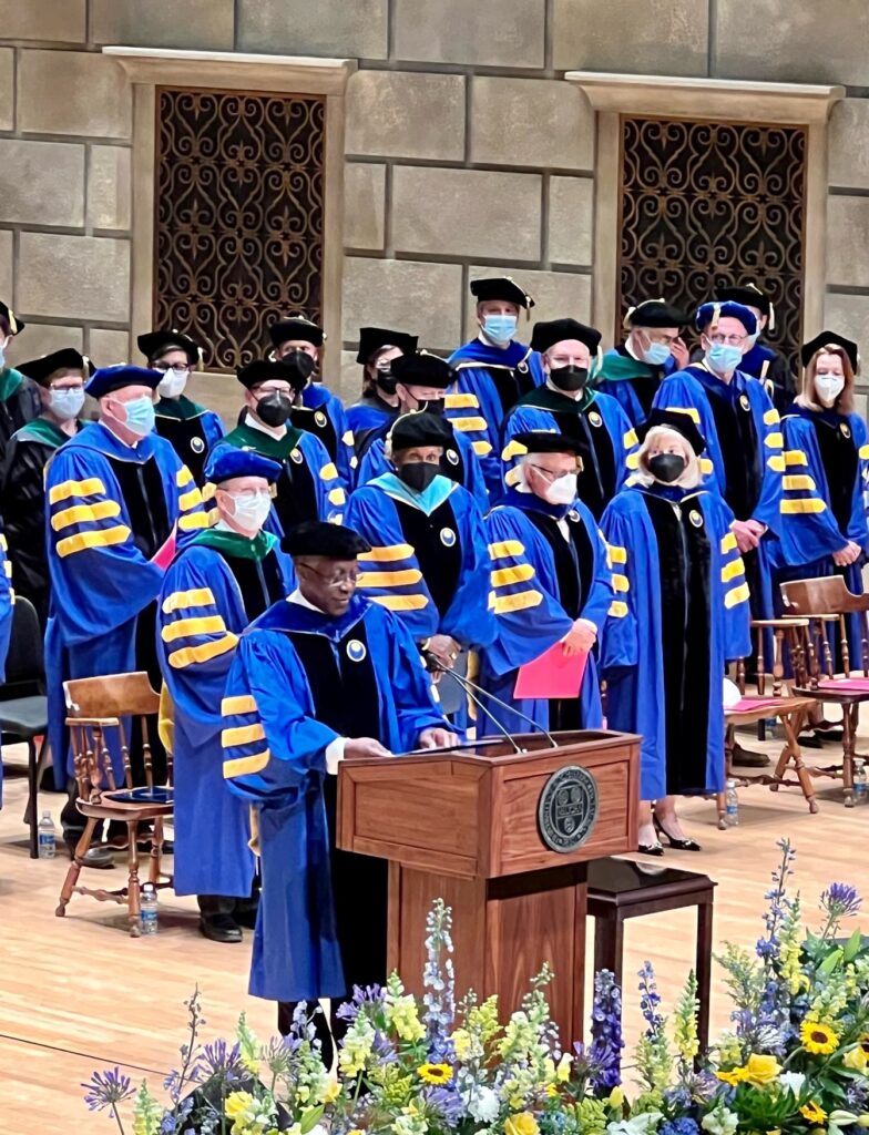A dozen adults wear royal blue gowns standing behind the podium at a graduation ceremony