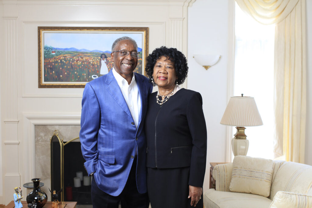 A man in a blue suit and a woman in a black suit stand in a living room in front of a painting.