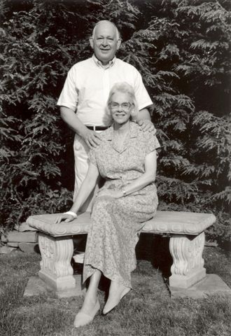 A black and white photo of a woman in a floral dress sitting on a stone bench with a man in a white shirt standing with his hand on her shoulder.