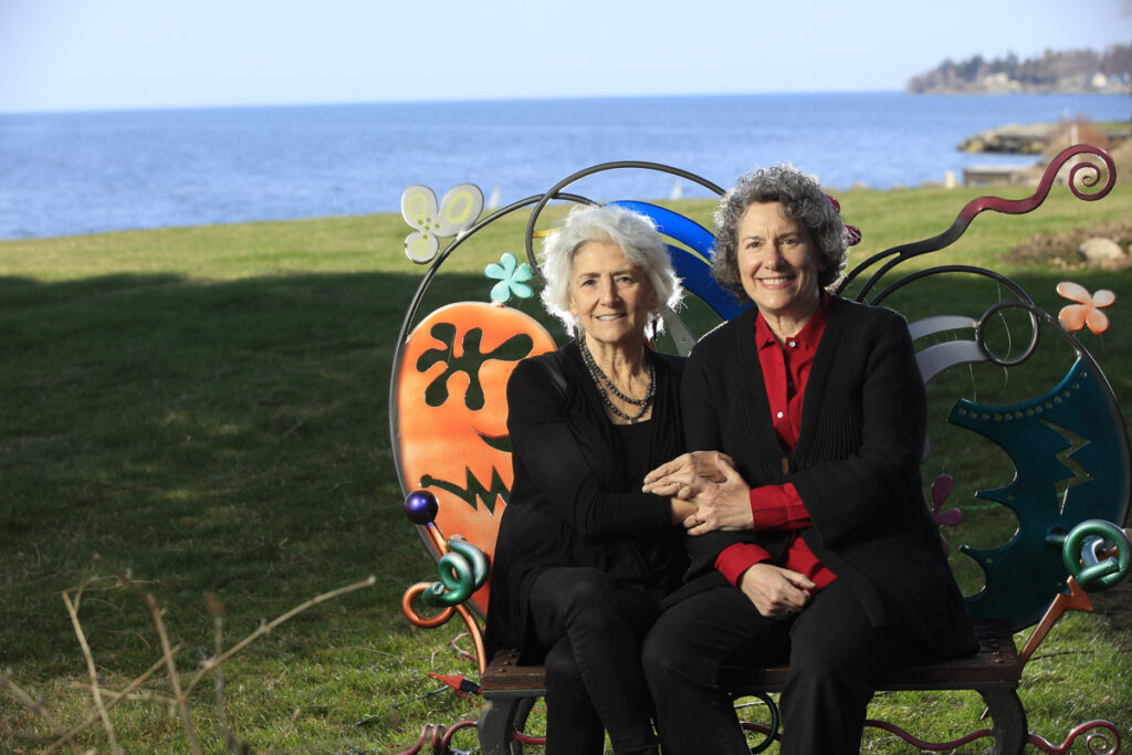 Two people sit on an artsy decorated bench with a green field and a lake in the background
