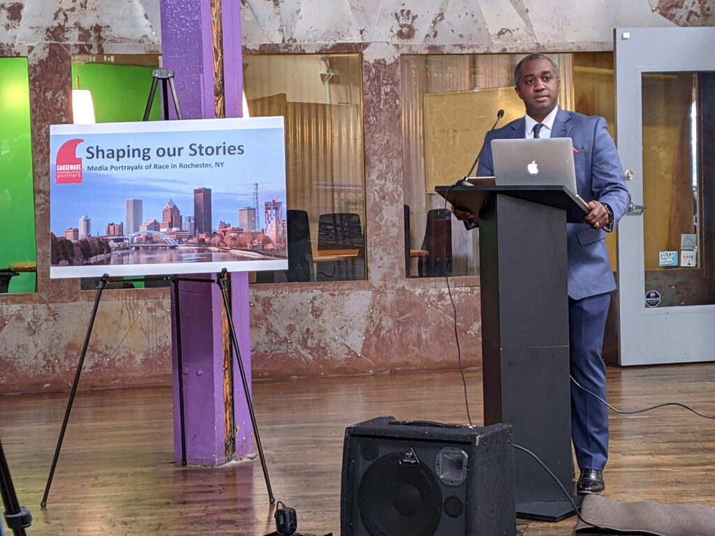 A man stands at a podium next to a poster that says Shaping our Stories: media portrayals of race in Rochester, NY