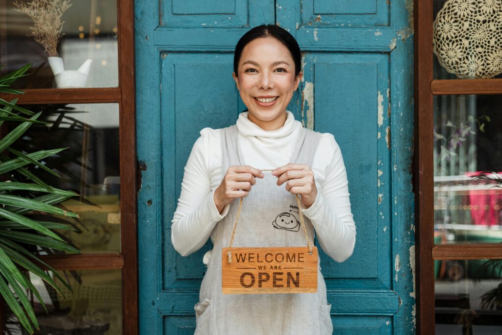 Shop owner stands outside her blue door with a "Welcome we are open" sign