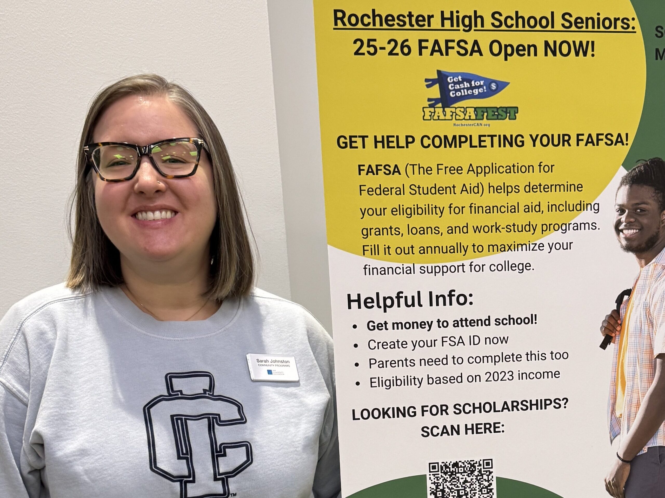 A girl in an Ithaca College sweatshirt stands next to a FAFSA Fest poster