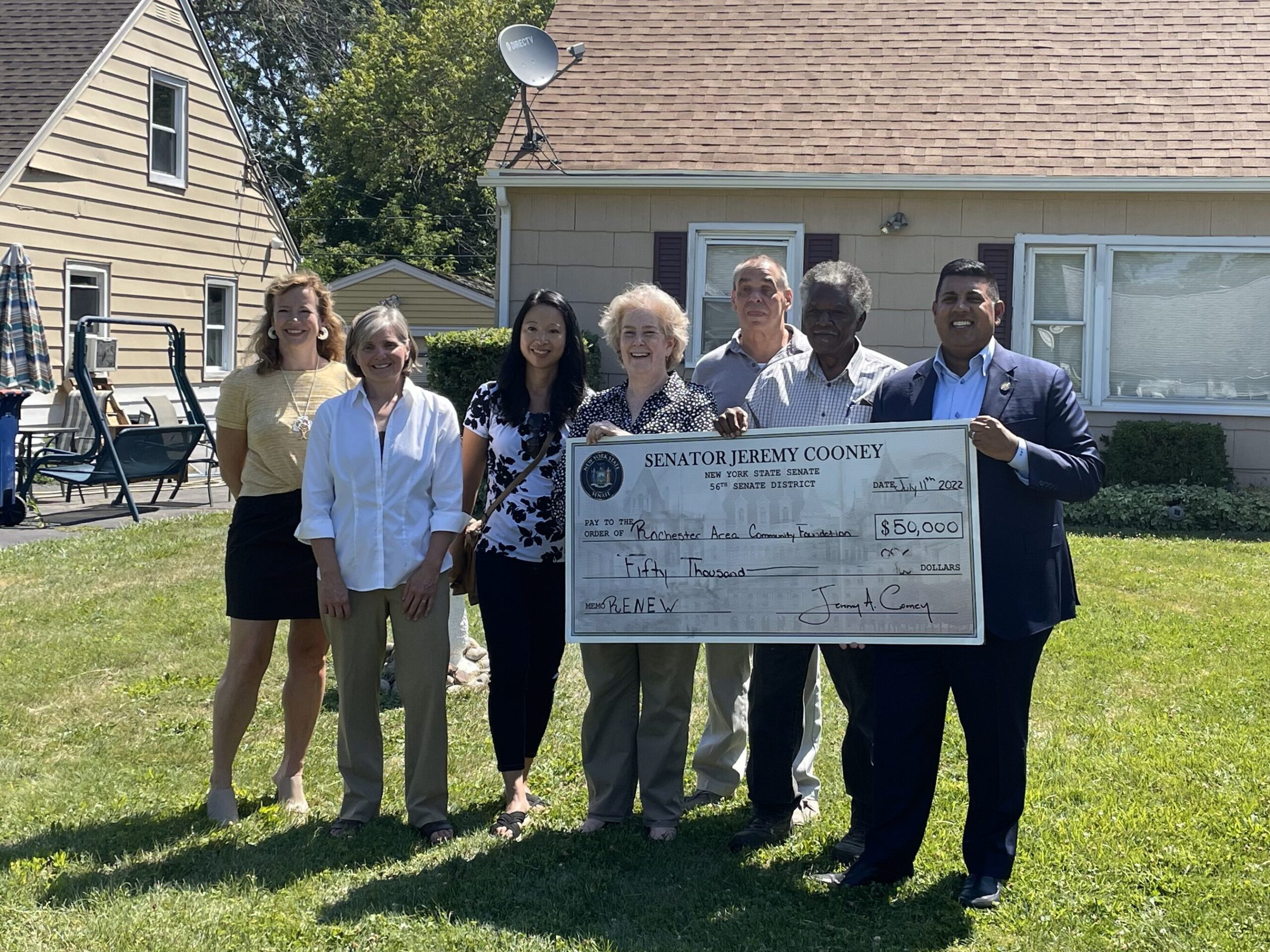 Seven people hold a giant check for $50,000 for RENEW on the front lawn of a house.