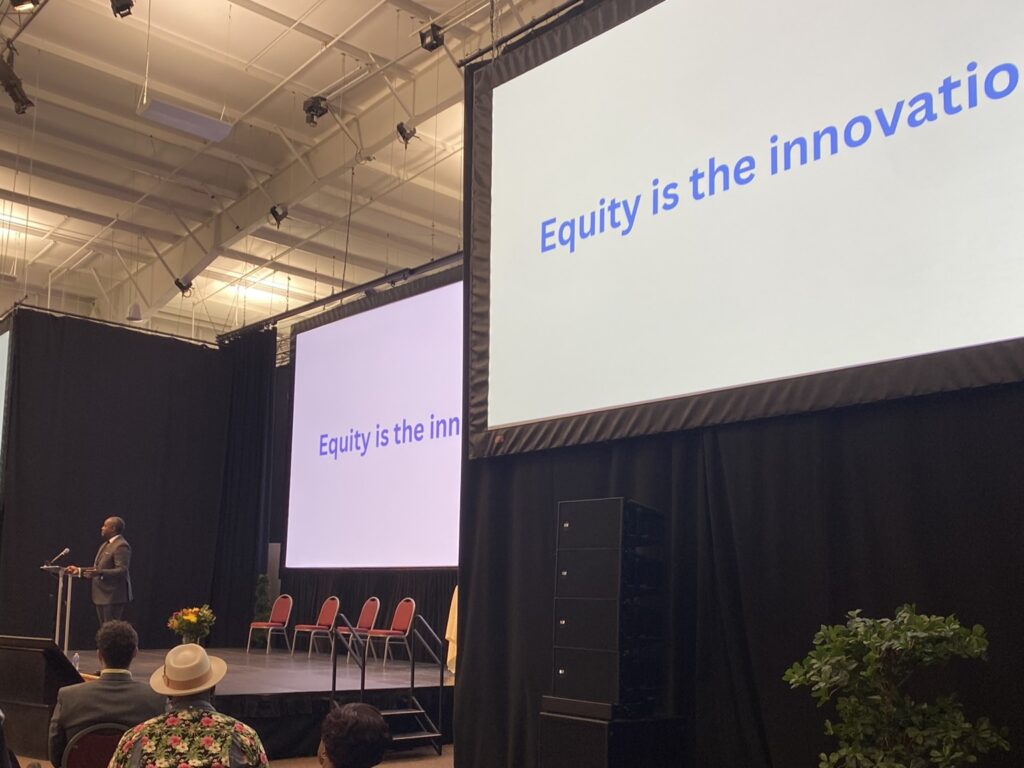 The words "equity is in the innovation" on screen behind a man speaking at a podium.