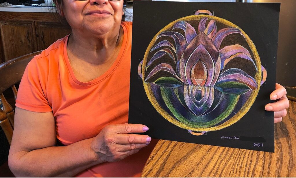 A woman in an orange shirt holds up her drawing: a gold circle with a purple and green flower