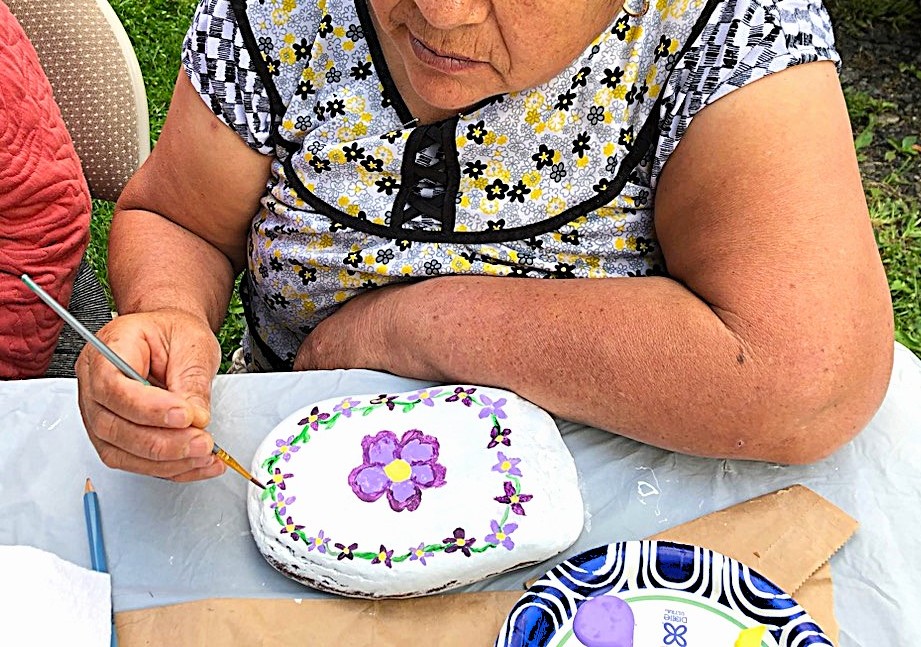 A woman in a grey and yellow shirt paints colorful flowers on a rock