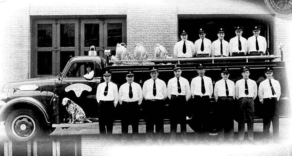 A vintage photo of fifteen men in white shirts and stand in front of a firetruck with a dalmatian.