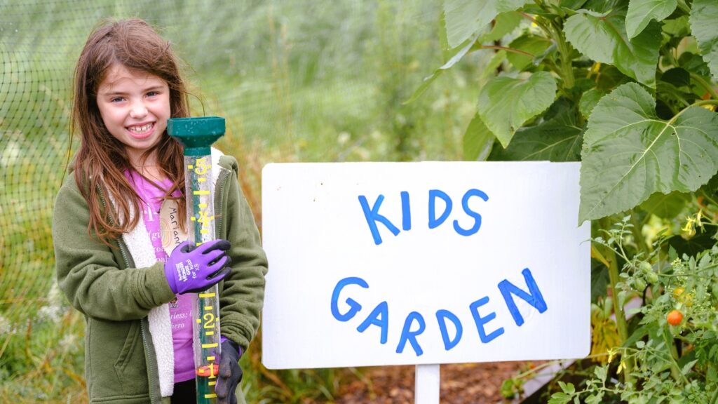 A young girl holds a rain meter next to a sign that says Kids Garden