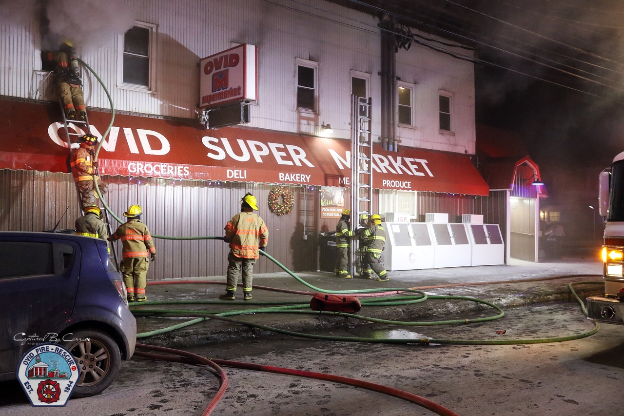 Firefighters climbing up a ladder to a window filled with smoke in front of Ovid Super Market
