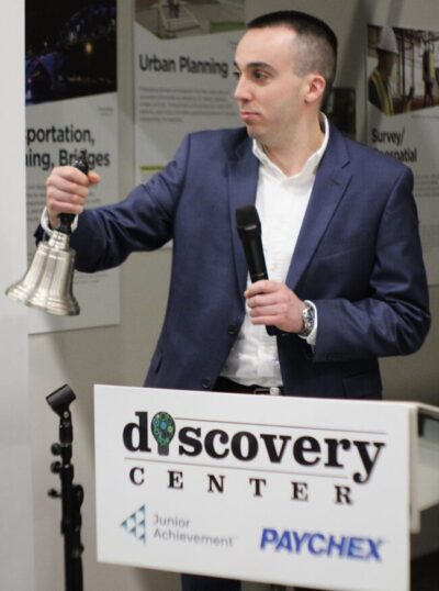 A man ringing a bell and holding a microphone in front of a sign that says Discovery Center with the Junior Achievement and Paychex logos