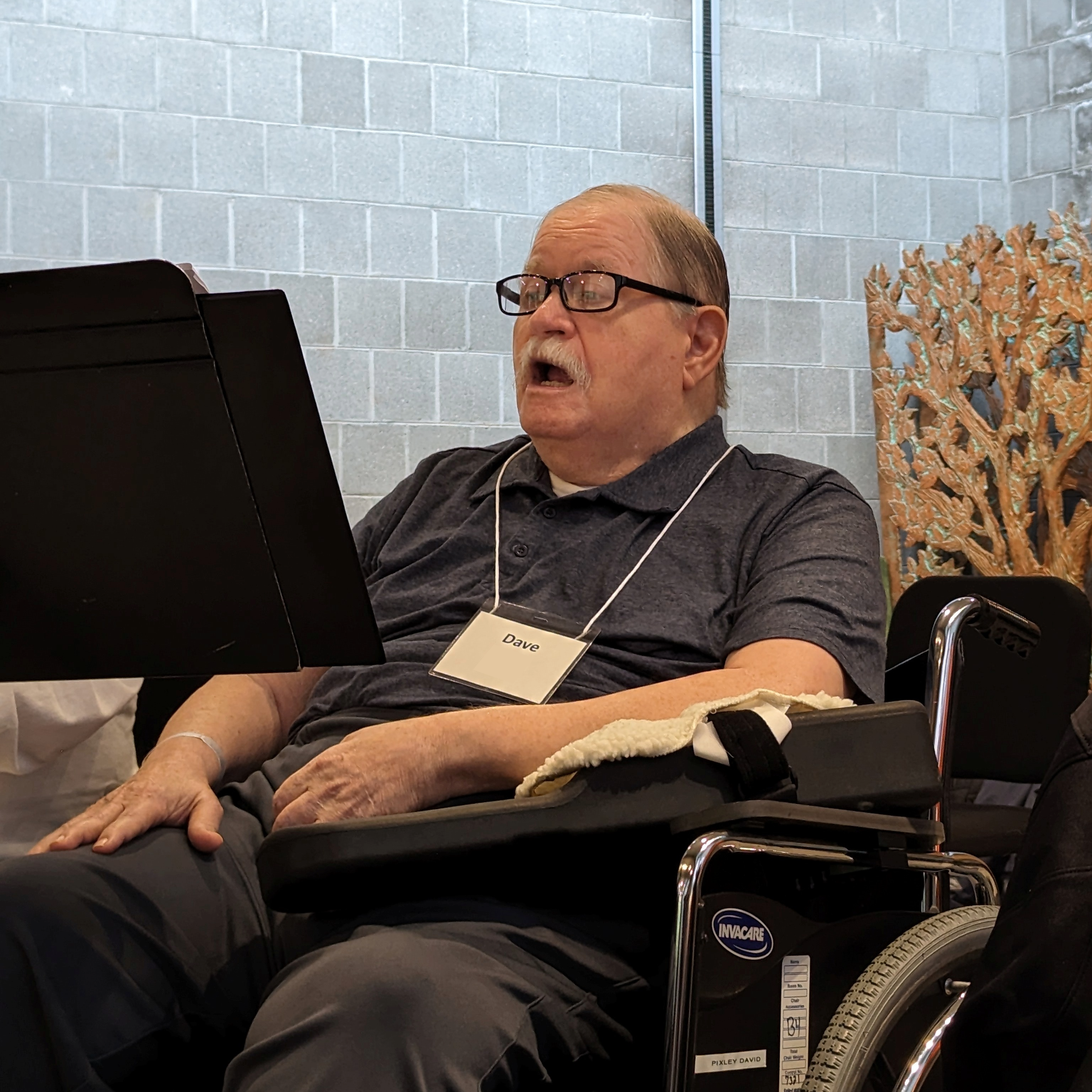 A man in a wheelchair singing in front of a gray brick wall.