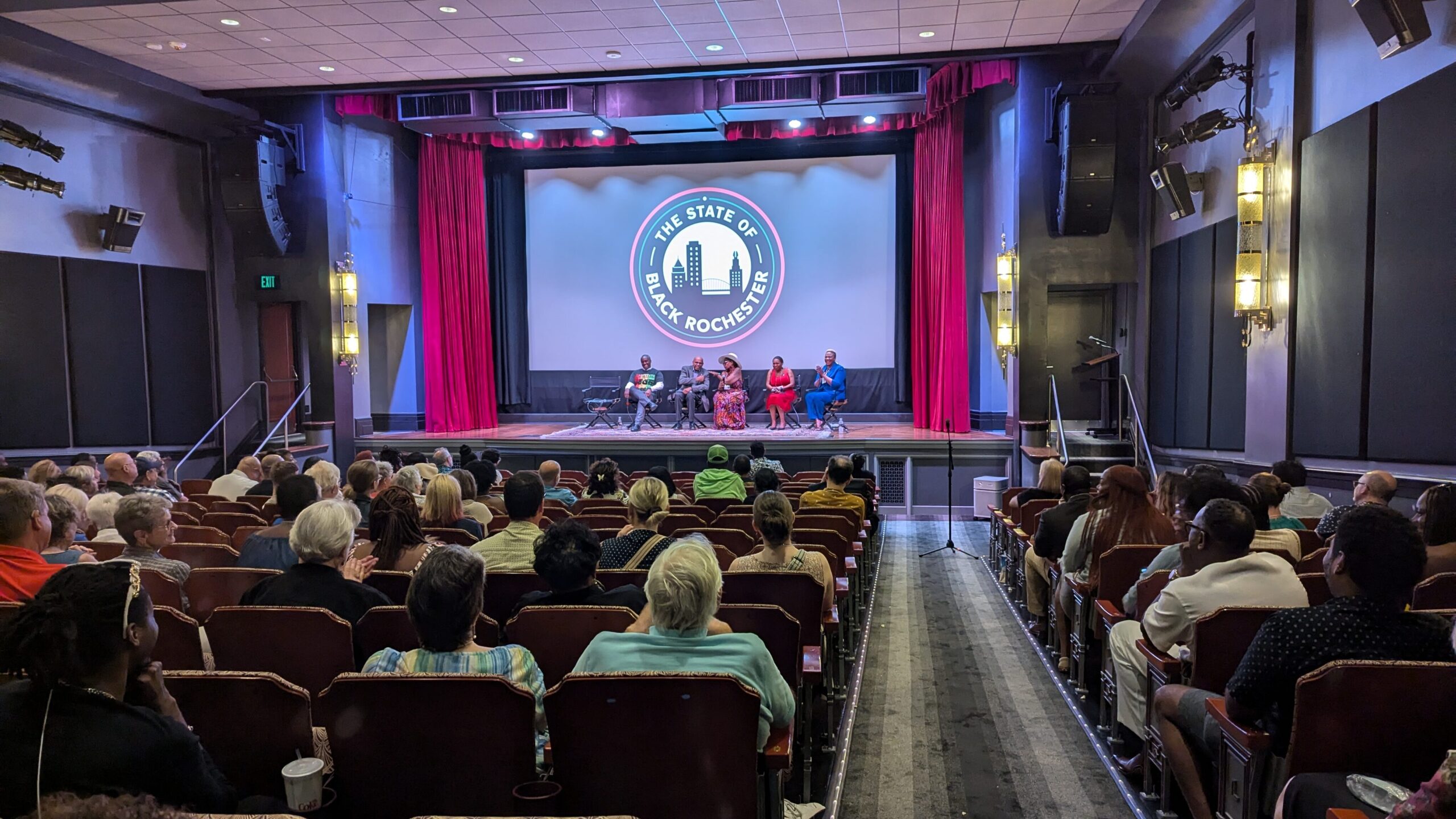A theatre full of people look at the stage with five panelists and a screen that reads "The State of Black Rochester."