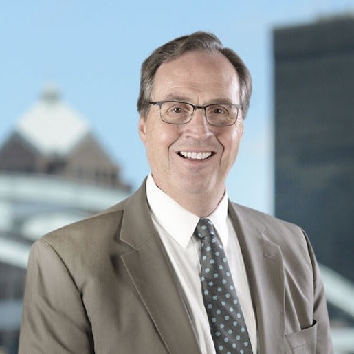A man in a beige suit and glasses stands in front of the Rochester city skyline
