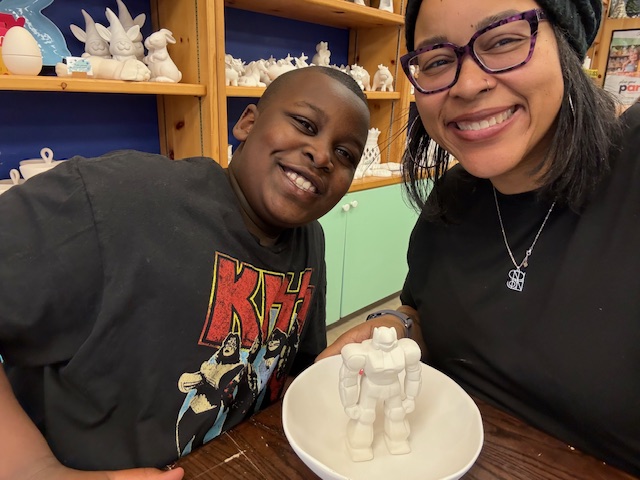 A woman in purple glasses and a young boy make an action figure out of clay.