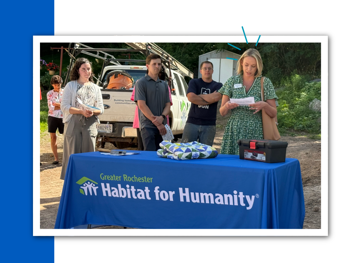 A woman in a green dress reads a speech in front of a Greater Rochester Habitat for Humanity table