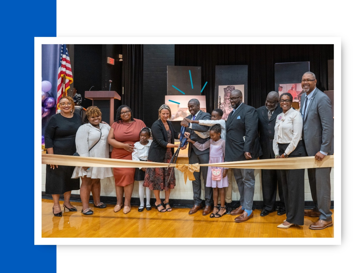 A man in a gray suit holds giant scissors in a line of people cutting a yellow ribbon,