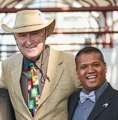 A man in a beige hat and suit and cartoon tie next to a man in a purple bowtie.