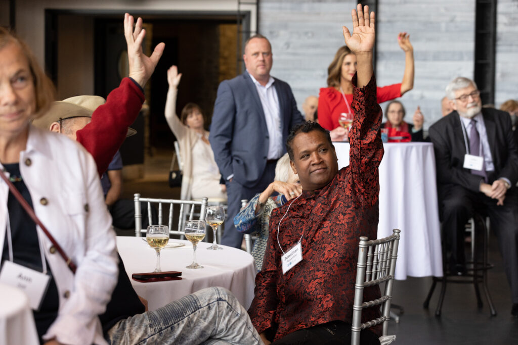 A man in a red and black formal shirt raises his hand at a gala.