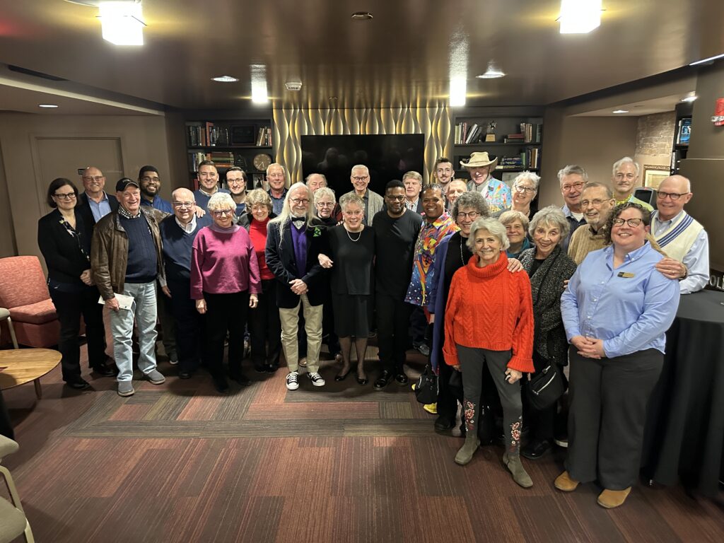 A large group shot of people in bright clothing in a room with maroon carpet and a gold fireplace.