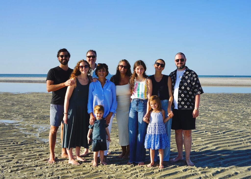 A family group shot with three generations on a beach in front of a blue sky.