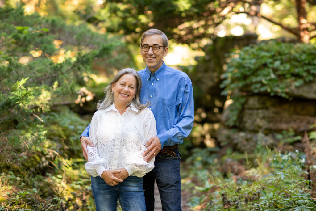 Outdoors, a man with glasses and a blue button-down shirt rests his hands on a woman's shoulders who wears a loose white lace top.