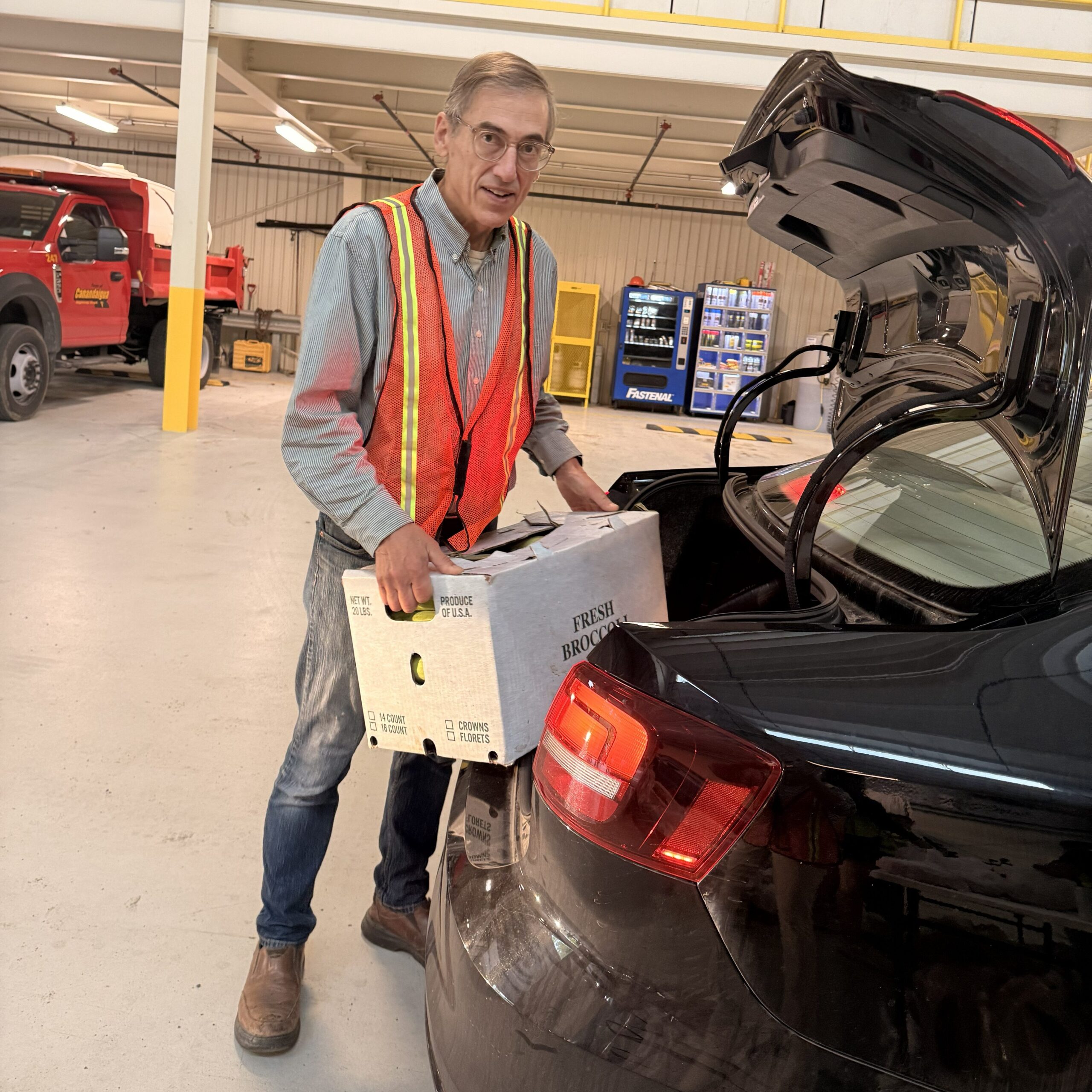 A man in an orange safety vest unloads a box from the back of a car.