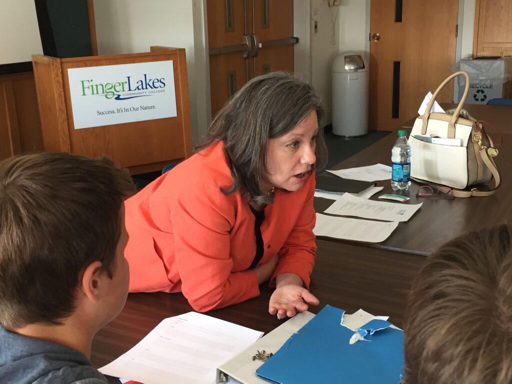 A woman in an orange blazer leans across a desk talking to people in front of a podium sign that says Finger Lakes Community College.