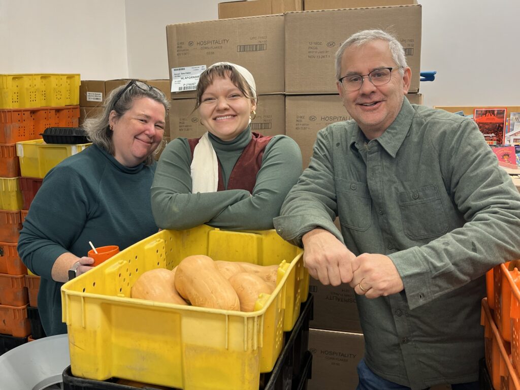 Three people smile in front of a bucket of vegetables.