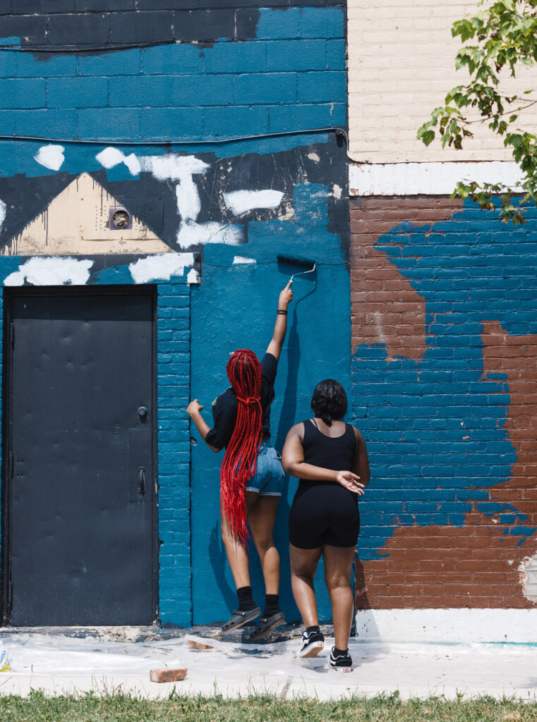 Two young people paint old, multi-colored brick walls dark teal with a roller.