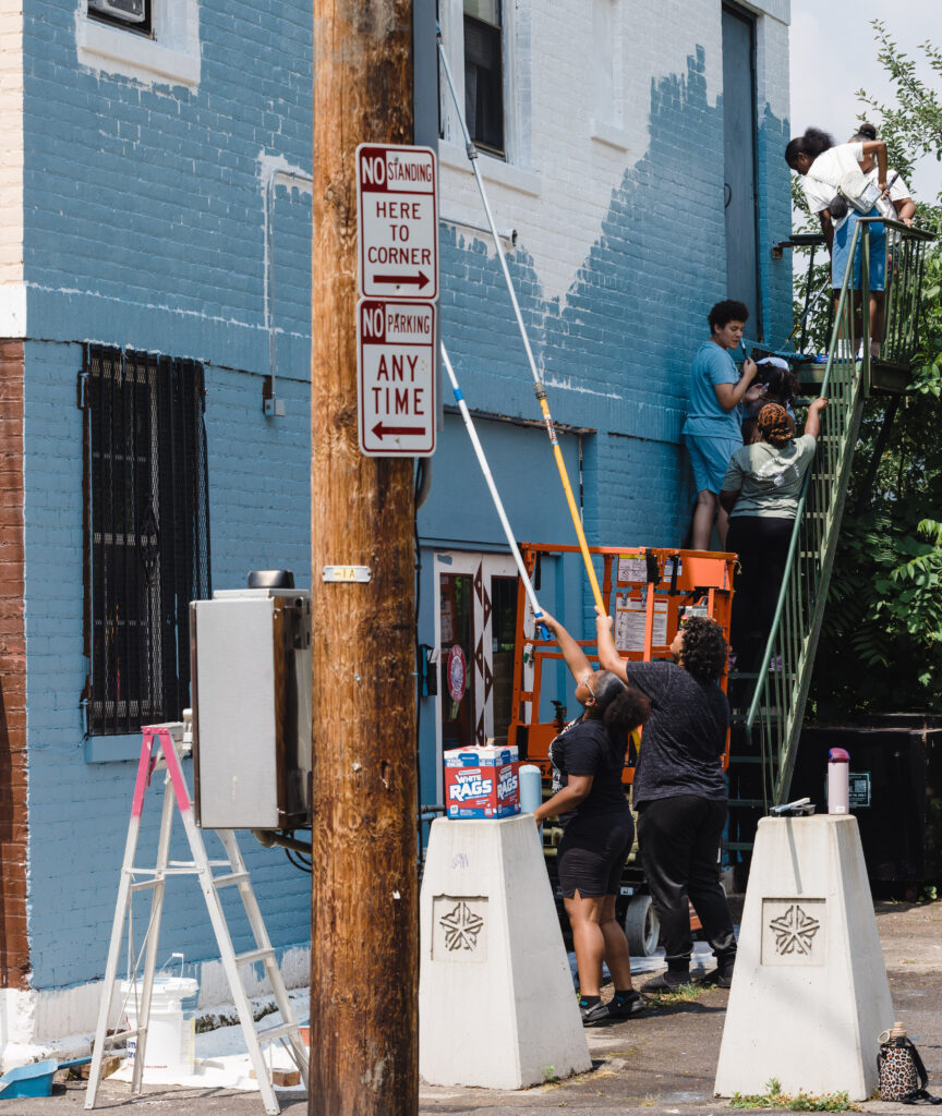 Six young people painting a two story commercial building blue with long rollers