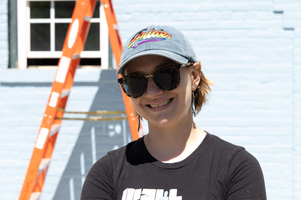 A person in a blue pride baseball had in sunglasses in front of a ladder.
