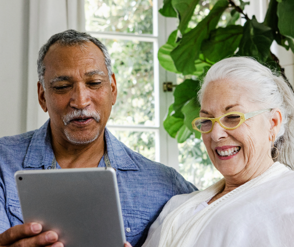 An older man holds on ipad sitting indoors next to a woman smiling in yellow glasses.